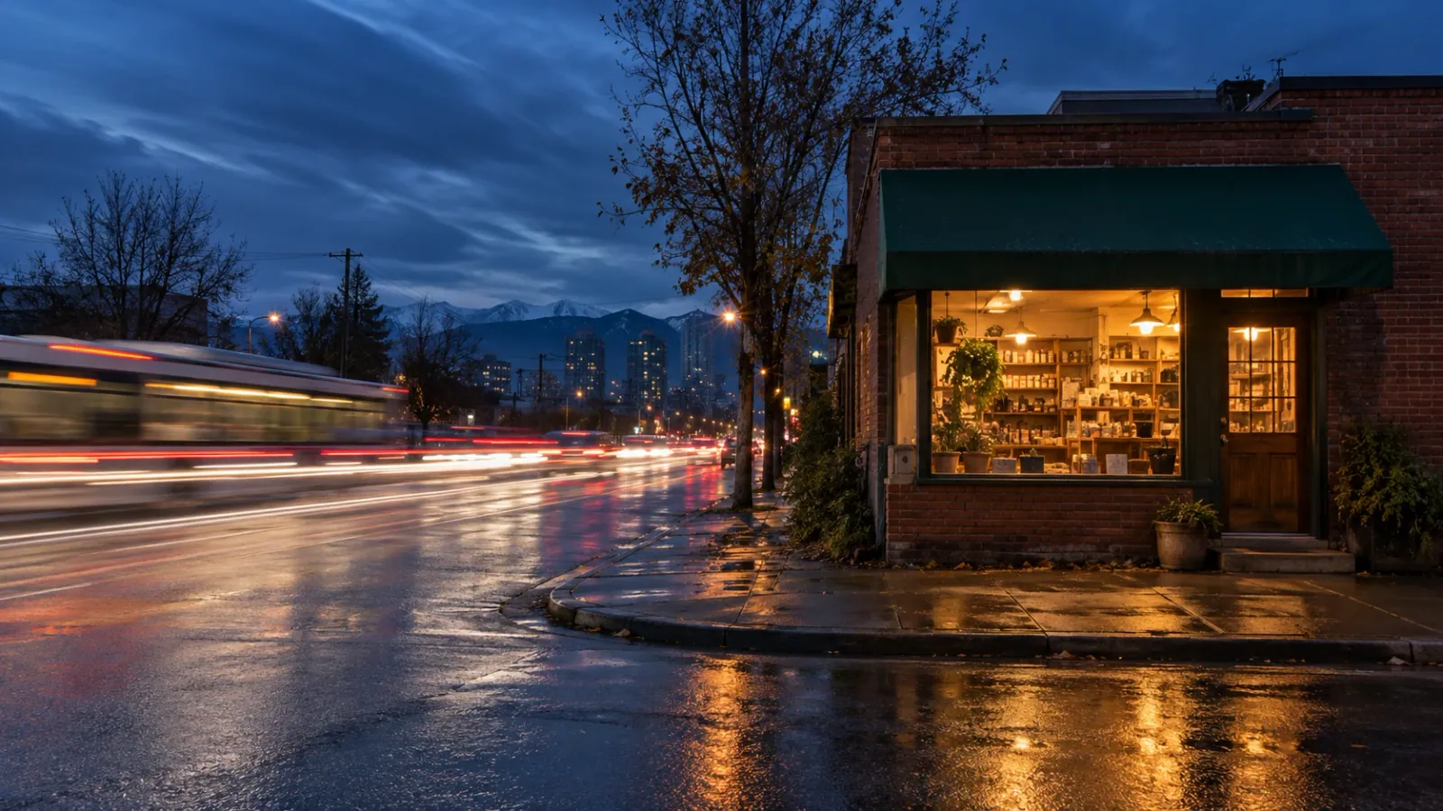 A small brick shop with a green awning and a warmly lit window on a wet street corner at dusk, with traffic blurred into light trails beside it